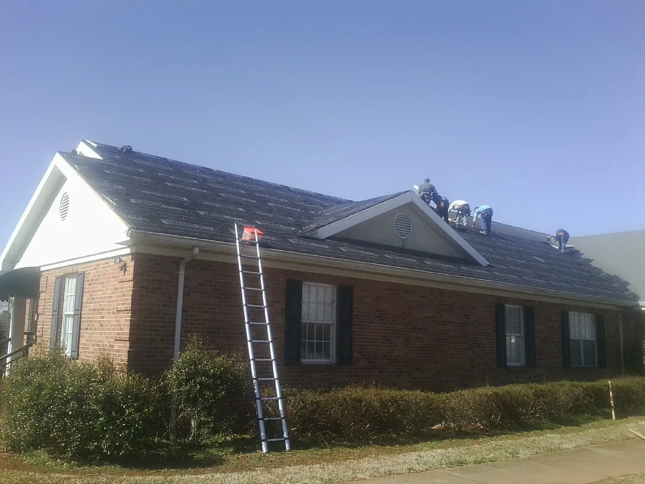 Crew tearing shingles off a brick ranch roof with a ladder set — Emergency Roof Repair work by our crew in West Eaton