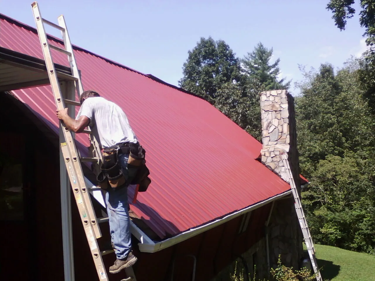 Roofer on a ladder beside a red metal roof and stone chimney — Soffit & Fascia Repair by Roofing Brothers in West Eaton